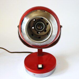 May include: A vintage red and chrome desk lamp with a round head and base. The lamp features a silver-colored rim around the light and a small on/off switch. A brown power cord extends from the base.