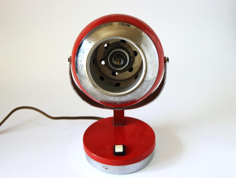 May include: A vintage red and chrome desk lamp with a round head and base. The lamp features a silver-colored rim around the light and a small on/off switch. A brown power cord extends from the base.