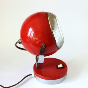 May include: A vintage red desk lamp with a spherical head and a chrome-colored trim. The lamp has a round base with a switch and a power cord. The lamp is made of metal and has a retro design.