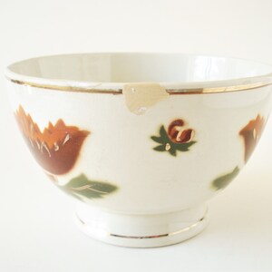 May include: A white ceramic bowl with a gold rim and floral design. The bowl features brown and orange flowers with green leaves.