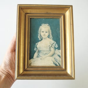May include: A framed portrait of a young girl with blonde curly hair, wearing a light-colored dress. The portrait is set against a teal background and is enclosed in a gold-colored wooden frame.