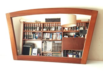 May include: A large, wooden-framed mirror with a unique, asymmetrical shape. The mirror reflects a room with bookshelves, a lamp, and various objects. The frame is a warm, reddish-brown color, adding a touch of elegance to the space.