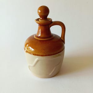 Può includere: Una piccola brocca in ceramica marrone e bianca con un manico rotondo e un pomo sul coperchio.