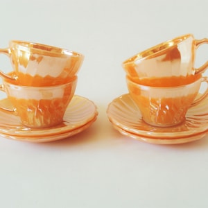 Può includere: Sei tazze e piattini arancioni. Le tazze sono impilate a coppie, ciascuna con un manico e un esterno strutturato. I piattini hanno un bordo smerlato. Il set è esposto su una superficie bianca.