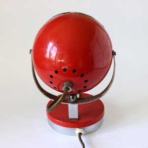 May include: A vintage red desk lamp with a spherical shade and a circular base. The lamp has a metal frame and a power cord. The shade has small ventilation holes. The lamp is a retro design.