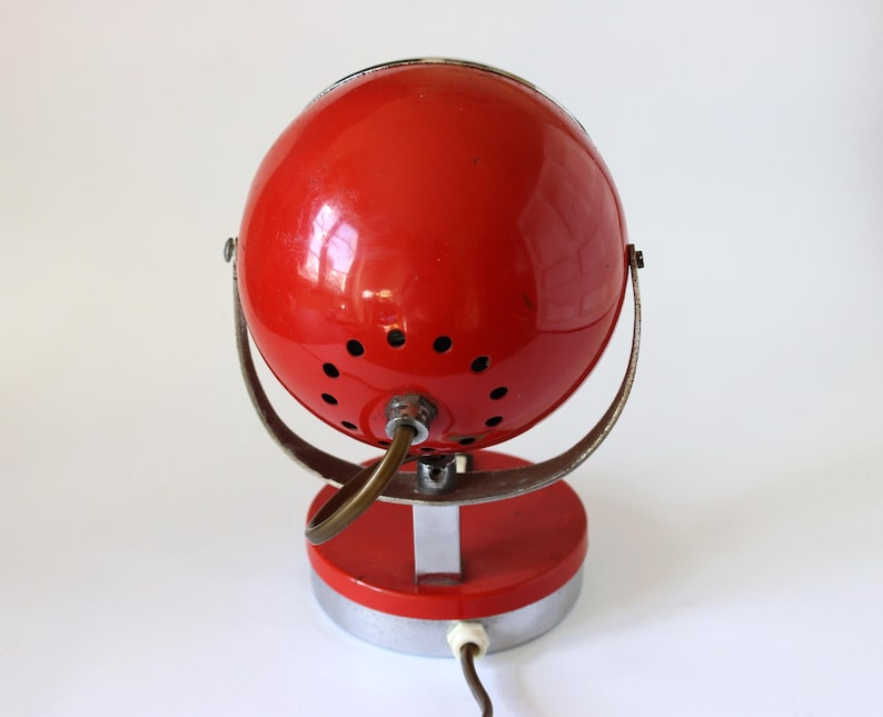 May include: A vintage red desk lamp with a spherical shade and a circular base. The lamp has a metal frame and a power cord. The shade has small ventilation holes. The lamp is a retro design.