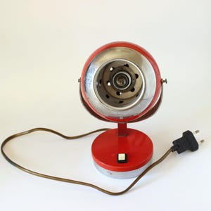 May include: A vintage red and silver desk lamp with a round head and base. The lamp has a silver rim around the light and a brown power cord with a black plug. The lamp has a small white button on the base.