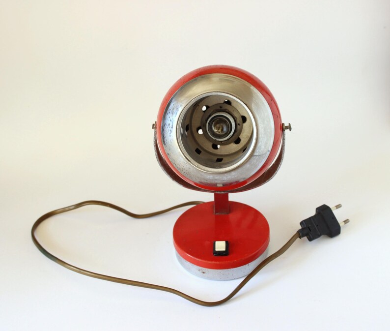 May include: A vintage red and silver desk lamp with a round head and base. The lamp has a silver rim around the light and a brown power cord with a black plug. The lamp has a small white button on the base.