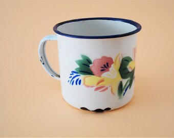 May include: White enamel mug with a blue rim and a floral design in pink, yellow, green, and blue.