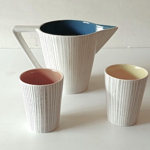 Puede incluir: Una jarra de cerámica blanca con interior azul y asa blanca, acompañada de dos tazas a juego. La jarra y las tazas presentan un patrón de rayas verticales grises. Una taza tiene el interior rosa, la otra amarillo.