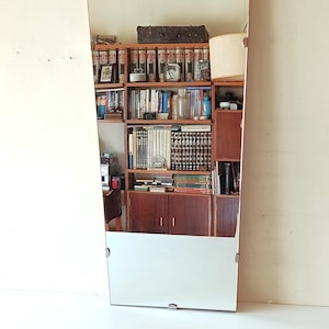 Puede incluir: Un espejo alto con la parte superior arqueada y forma rectangular. El espejo refleja una estantería de madera llena de libros, una lámpara y una maleta. El espejo está montado en una pared blanca con herrajes plateados.