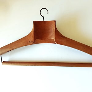 May include: A vintage wooden clothes hanger with a dark metal hook and a wooden bar. The hanger is made of light brown wood and has a wide, curved design. The hanger is designed to hold clothing.