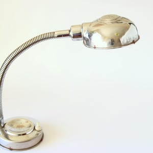 Puede incluir: Una lámpara de escritorio plateada con un brazo flexible de cuello de cisne y una pantalla redonda en forma de cúpula.