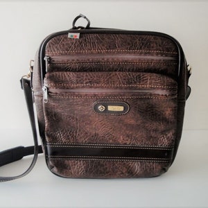 May include: A brown leather shoulder bag with multiple zippered compartments and a long adjustable strap. The bag features a textured surface, a small logo, and a top handle. The bag has a rectangular shape and is designed for carrying items.