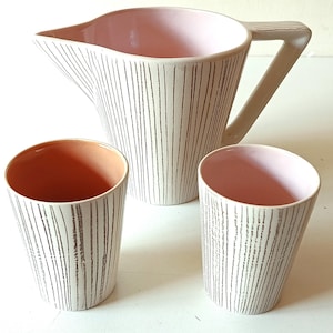 Puede incluir: Una jarra de cerámica y dos vasos a juego. El conjunto presenta un exterior color crema con rayas verticales marrones. El interior de la jarra y los vasos es de color rosa claro. La jarra tiene un asa triangular.