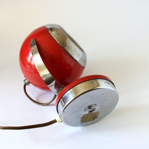 May include: A vintage red and silver desk lamp with a spherical shade and a circular base. The lamp features a curved metal arm and a silver accent on the shade. The lamp is shown on a white background.