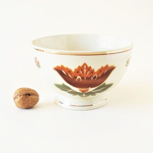 May include: A white ceramic bowl with a brown and green floral design. The bowl has a gold rim and a slightly flared lip.