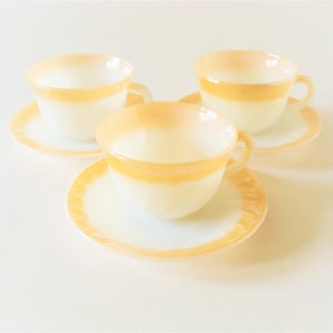 以下が含まれることがあります： 3つのヴィンテージティーカップとソーサー。カップとソーサーは白で、カップの上部とソーサーの縁にオレンジ色の帯があります。カップには取っ手が付いています。