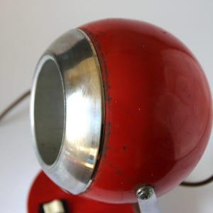 May include: A vintage red desk lamp with a spherical head and a silver-colored trim. The lamp has a small, round base and a curved metal handle. The lamp's design is retro and functional.