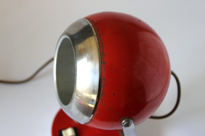May include: A vintage red desk lamp with a spherical head and a silver-colored trim. The lamp has a small, round base and a curved metal handle. The lamp's design is retro and functional.
