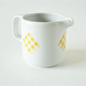 May include: White ceramic milk jug with a yellow gingham pattern. The jug has a handle and a spout.
