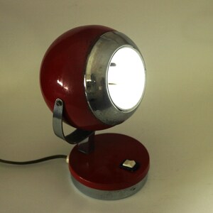 May include: A vintage red and silver desk lamp with a spherical head and a circular base. The lamp has a bright, illuminated light and a switch on the base. The lamp is connected to a black power cord.