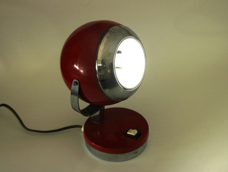May include: A vintage red and silver desk lamp with a spherical head and a circular base. The lamp has a bright, illuminated light and a switch on the base. The lamp is connected to a black power cord.