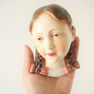 May include: A vintage porcelain figurine of a girl with brown braids and pink bows. The figurine has a white dress and a painted face.