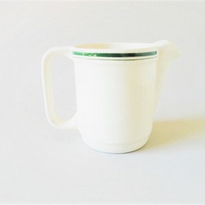 May include: Cream-colored ceramic creamer with a handle and spout. A thin green stripe encircles the top rim. The creamer is set against a plain white background, highlighting its simple, functional design.