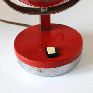 May include: A vintage red and silver desk lamp with a circular base and a push button switch. The lamp has a red shade and a silver metal frame. The lamp's base is approximately 4 inches in diameter.