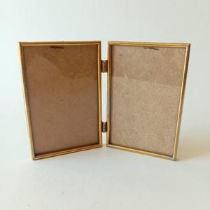 Puede incluir: Un marco de fotos de metal dorado con dos aberturas. El marco está abierto y listo para mostrar fotos.