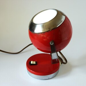 May include: A vintage red and chrome desk lamp with a spherical head and a circular base. The lamp has a silver trim around the light opening and a small on/off switch. A brown cord extends from the base.