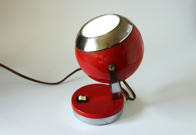 May include: A vintage red and chrome desk lamp with a spherical head and a circular base. The lamp has a silver trim around the light opening and a small on/off switch. A brown cord extends from the base.
