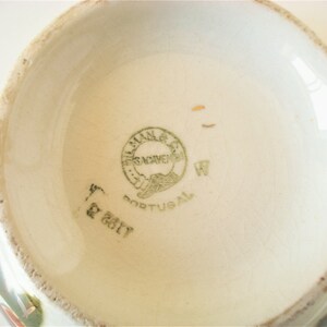 May include: Close-up of the bottom of a ceramic bowl with a green and white floral design. The bowl has a green stamp that reads "F&aacute;brica de Novemeis, Portugal" and a handwritten "W" next to it.