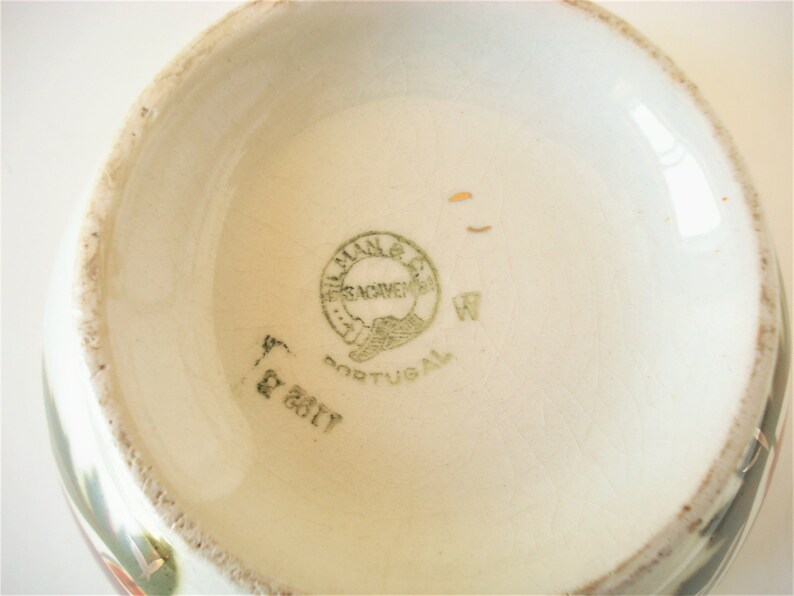 May include: Close-up of the bottom of a ceramic bowl with a green and white floral design. The bowl has a green stamp that reads "F&aacute;brica de Novemeis, Portugal" and a handwritten "W" next to it.