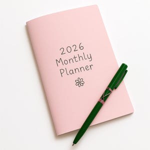 Pode incluir: Um planejador mensal rosa para 2026 com uma caneta verde em cima. O planejador tem um pequeno desenho de flor na frente.