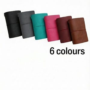 May include: Six leather journals in various colors: black, gray, teal, pink, burgundy, and brown. Each journal is closed with an elastic band. The text "6 colours" is displayed below the journals.