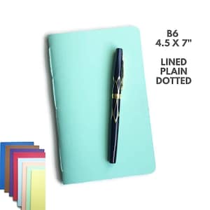 Puede incluir: Un cuaderno azul claro con un bolígrafo negro en la parte superior. El cuaderno mide 4,5 x 7 pulgadas y tiene las opciones de papel rayado, liso o punteado. El cuaderno forma parte de un conjunto de cuadernos de varios colores.
