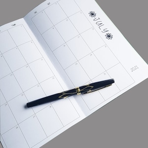 Pode incluir: Um planejador de calend&aacute;rio mensal em branco com uma caneta preta em cima. O calend&aacute;rio est&aacute; aberto para julho de 2021.