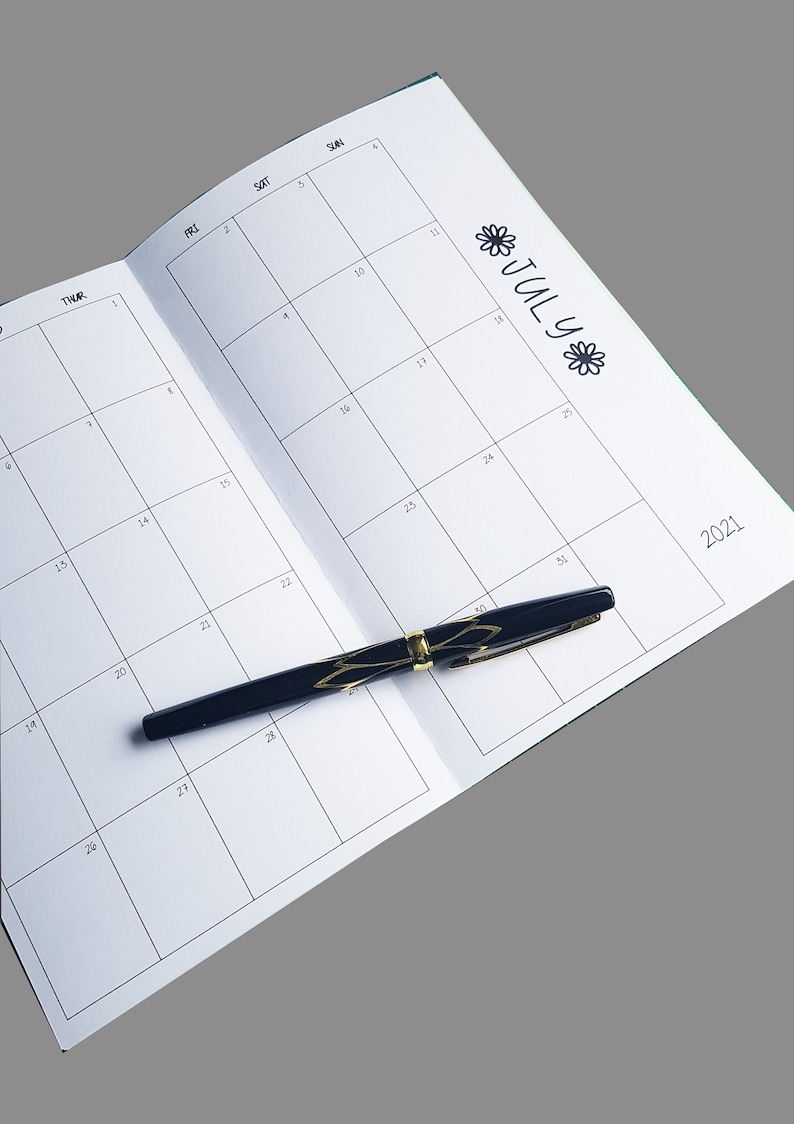 Pode incluir: Um planejador de calend&aacute;rio mensal em branco com uma caneta preta em cima. O calend&aacute;rio est&aacute; aberto para julho de 2021.