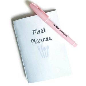 Peut inclure: Un carnet de planification des repas blanc avec le texte "Meal Planner" et une image d'une fourchette, d'un couteau et d'une cuillère. Un surligneur rose avec le texte "Wilko Highlighter" est posé sur le carnet.