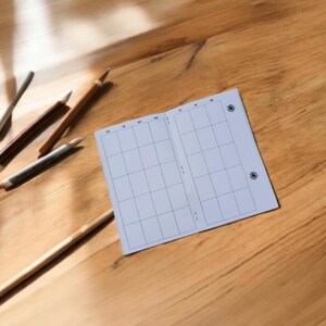 Pode incluir: Um planejador branco, em branco, quadriculado com um pequeno desenho de flor no canto superior direito. O planejador est&aacute; aberto para uma semana.