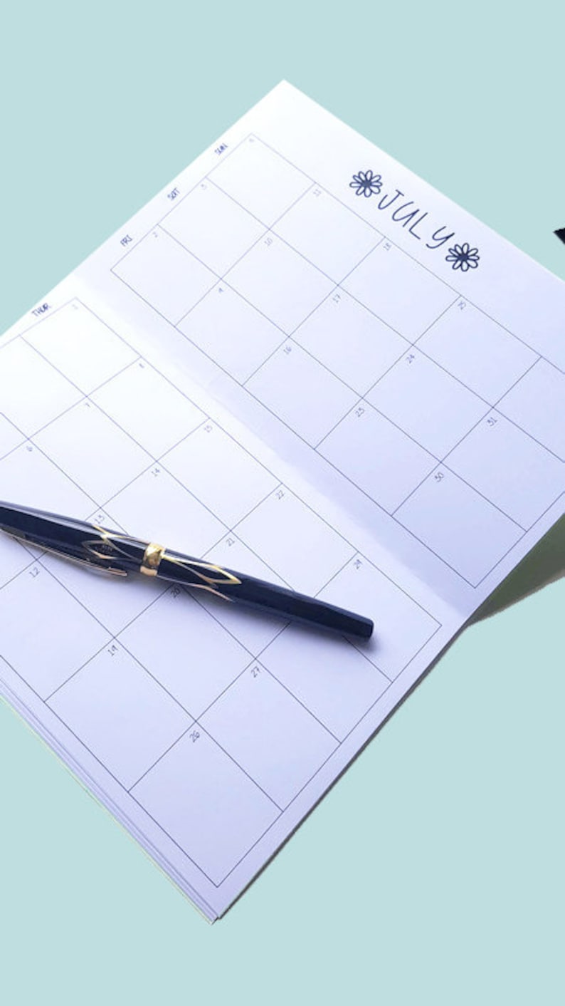 Pode incluir: Um calend&aacute;rio mensal branco com a palavra "Julho" escrita em tinta preta e decorado com pequenos desenhos de flores. Uma caneta preta com detalhes dourados est&aacute; sobre o calend&aacute;rio.