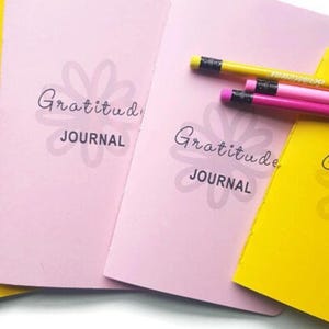 Puede incluir: Cuatro diarios de gratitud, dos amarillos y dos rosas, con el texto "Gratitude Journal" y un diseño de flor estilizado. Los diarios están apilados uno encima del otro y hay tres lápices encima de los diarios.