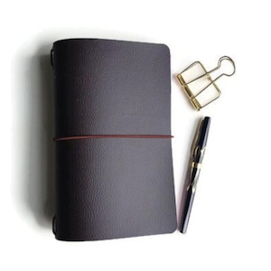 May include: A dark brown leather bound journal with a brown elastic band closure. A gold binder clip and a black pen are on a white surface next to the journal.