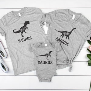 May include: Three gray t-shirts with dinosaur silhouettes and the words "Daddy Saurus", "Mommy Saurus", and "Baby Saurus" printed on them.