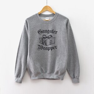 Puede incluir: Una sudadera gris con el texto "Gangster Wrapper" en letras negras. Una ilustración en blanco y negro de una caja de regalo envuelta con un lazo está por encima del texto.