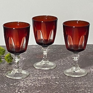 May include: Three matching stemmed glasses featuring deep red bowls with vertical panel cuts and clear glass stems and circular bases. The glasses have a rounded shape and a smooth finish, showcasing a consistent design across all three pieces.