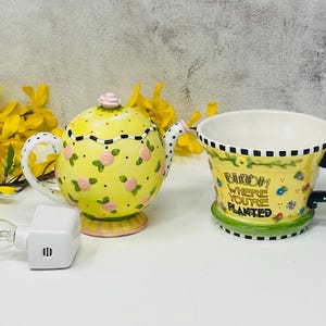 Puede incluir: Una tetera de cerámica amarilla con detalles florales rosas y una taza a juego con el texto "Bloom Where You're Planted". También hay una luz de noche blanca. La taza tiene un asa de lunares blancos y negros.