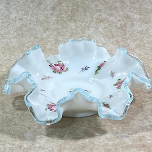 Pode incluir: Uma taça de vidro leitoso branco com uma borda ondulada e um acabamento azul claro. O interior da taça é decorado com um padrão de pequenas rosas cor-de-rosa e folhas verdes espalhadas pela superfície. A taça tem uma base circular.